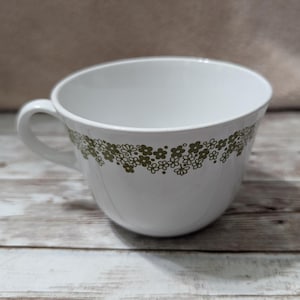 Może przedstawiać: Biały ceramiczny kubek do herbaty z zielonym wzorem kwiatowym wokół krawędzi. Kubek ma zaokrąglony kształt i uchwyt.
