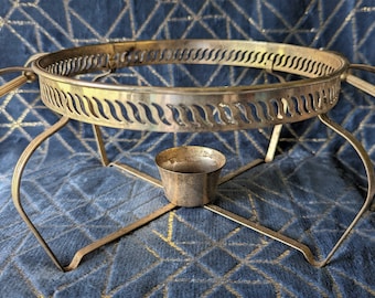 Pode incluir: Um suporte para chafing dish de metal dourado com uma parte superior redonda e três pernas. O suporte tem uma pequena taça redonda de metal no centro.
