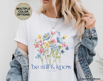 Könnte beinhalten: Ein weißes T-Shirt mit einer floralen Grafik im Aquarellstil, bestehend aus blauen, rosa und gelben Blumen, grünen Stielen und kleinen Schmetterlingen. Darunter steht der Text "be still & know" in blauer Schrift sowie "PSALM 46:10".