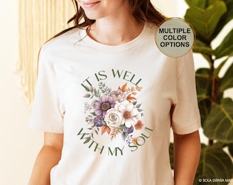 Puede incluir: Camiseta color crema con un diseño floral y el texto "IT IS WELL WITH MY SOUL" en verde. El arreglo floral incluye flores blancas, moradas y naranjas. Un botón circular en la camisa dice "MULTIPLE COLOR OPTIONS".
