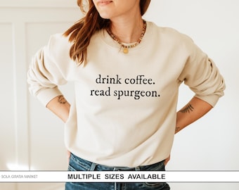 May include: A cream-coloured sweatshirt with the text "drink coffee. read spurgeon." in black. The long-sleeved top is worn with blue jeans. The sweatshirt is a casual style.
