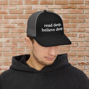 May include: A black and gray trucker hat featuring a structured front panel with the white text read deep. believe deep. printed in a clean sans-serif font. The cap includes a solid black brim and a breathable gray mesh back panel for ventilation.