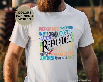 Op de afbeelding: Wit t-shirt met het woord "REFORMED" in een groot, gestileerd lettertype, omringd door andere woorden zoals "FORGIVEN" en "ADOPTED" in verschillende kleuren. Het shirt heeft ook de tekst "COLORS FOR MEN & WOMEN".
