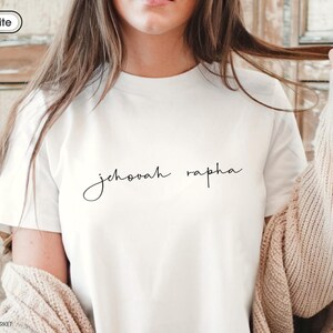May include: White t-shirt with the words "Jehovah Rapha" in a delicate, cursive font. The shirt is a classic crew neck style, made of a soft material. The text is centered on the chest.