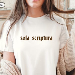 Puede incluir: Camiseta blanca con la frase "sola scriptura" en marrón con fuente gótica. La camiseta tiene cuello redondo y un diseño sencillo. El texto está centrado. La persona lleva un cárdigan beige.