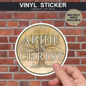 May include: Round vinyl sticker with the text "ABIDE IN CHRIST" and "John 15:4" in a stylized font, featuring a tree graphic. The sticker is tan and green, with the words in white. The background is a brick wall.