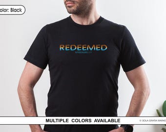 Op de afbeelding: Zwart T-shirt met het woord "REDEEMED" in een oranje en blauwe kleurovergang. De tekst "EPHESIANS 1:7" staat eronder. Het shirt met korte mouwen heeft de tekst gecentreerd op de borst.