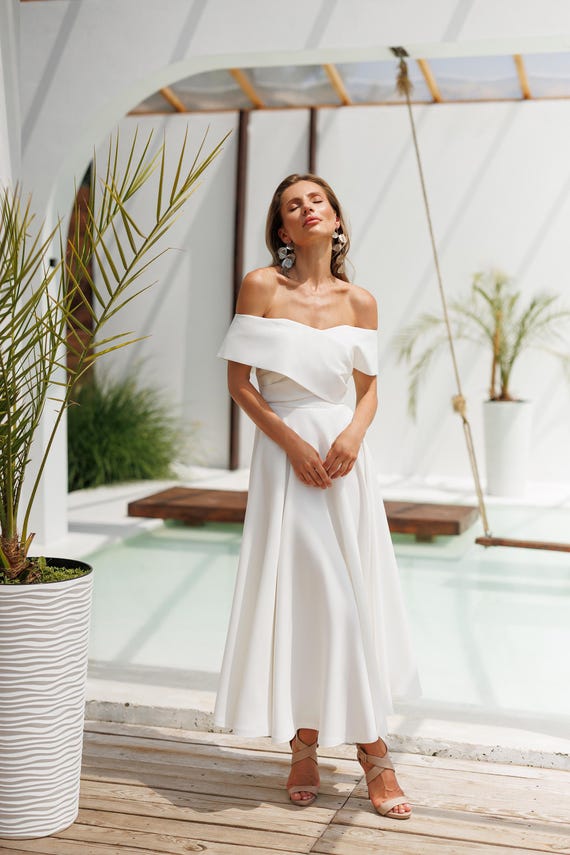 Elegant woman in an off-shoulder white summer dress, posing by a serene pool with greenery in the background.