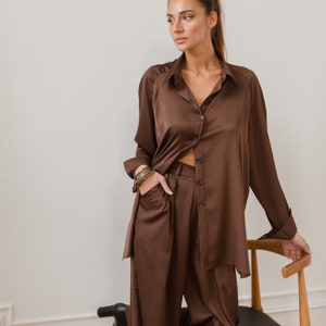 K&ouml;nnte beinhalten: Ein braunes Satin-Shirt und -Hosen-Set. Das Shirt hat einen Kragen und lange &Auml;rmel. Die Hose hat eine weite Beinweite und eine lockere Passform.