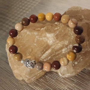 Puede incluir: Un brazalete de piedras multicolores con un dije de Buda plateado. El brazalete presenta un degradado de piedras marrones, amarillas y blancas.