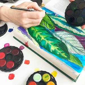 Op de afbeelding: Een persoon schildert een boek met aquarelverf. Het boek is open op een pagina met een tekening van groene bladeren. Er zijn twee ronde aquarelpaletten met verschillende kleuren verf.