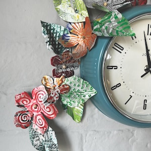 May include: A teal blue vintage-style clock with white face and black hands. The clock is decorated with a garland of colorful metal flowers and leaves.