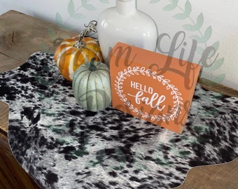 May include: A black and white cowhide table runner with a small orange pumpkin, a green pumpkin, and a wooden sign that says "Hello Fall" on a wooden table.