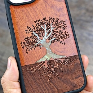 May include: A phone case with a wooden back featuring a tree design. The case has a dark brown border and a light brown wood grain back. The tree is carved with a silver-coloured trunk and roots.