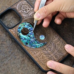 May include: A black phone case featuring a dark wood inlay with intricate laser-engraved patterns. A central yin-yang symbol is composed of iridescent blue and green abalone shell pieces, with a small circular abalone inlay in the dark wood section.
