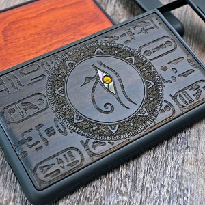 Op de afbeelding: Een zwarte telefoonhoes met een houten inlay met een Egyptisch oog van Horus-ontwerp. Het oog is omgeven door een cirkelvormig patroon en hi&euml;rogliefen.
