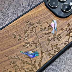 Könnte beinhalten: Eine Handyhülle aus Holz mit schwarzem Rand. Die Hülle hat ein geschnitztes Design eines Baumes mit zwei Vögeln aus irisierendem Perlmutt.