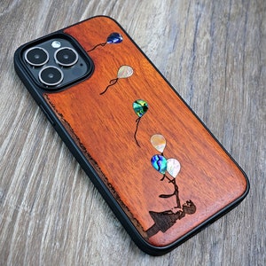 May include: A smartphone case with a black frame and a wood-grain back panel. The wood features a laser-engraved silhouette of a person holding strings attached to five iridescent, teardrop-shaped abalone shell inlays representing balloons.