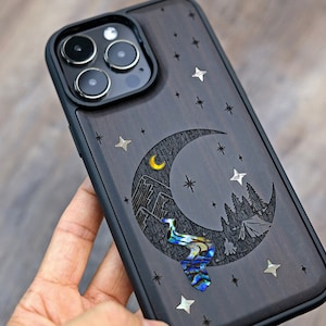 Puede incluir: Una funda para tel&eacute;fono de madera negra con un dise&ntilde;o de luna creciente. La luna est&aacute; tallada con una escena de monta&ntilde;a y un r&iacute;o hecho de concha de abul&oacute;n iridiscente. La funda est&aacute; decorada con peque&ntilde;as estrellas plateadas.
