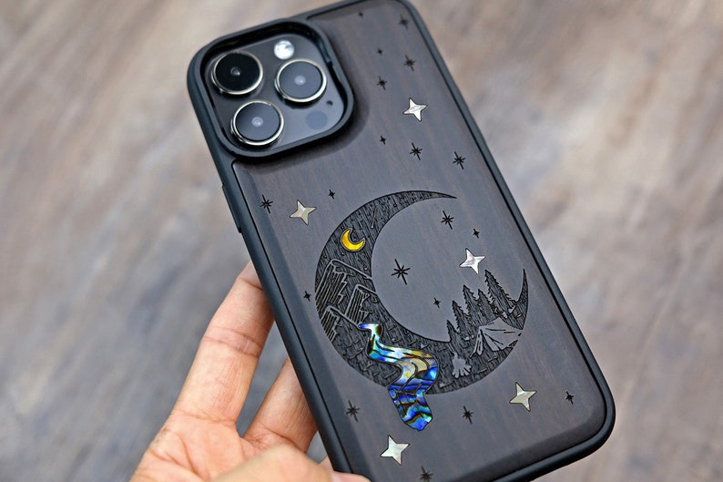 Puede incluir: Una funda para tel&eacute;fono de madera negra con un dise&ntilde;o de luna creciente. La luna est&aacute; tallada con una escena de monta&ntilde;a y un r&iacute;o hecho de concha de abul&oacute;n iridiscente. La funda est&aacute; decorada con peque&ntilde;as estrellas plateadas.