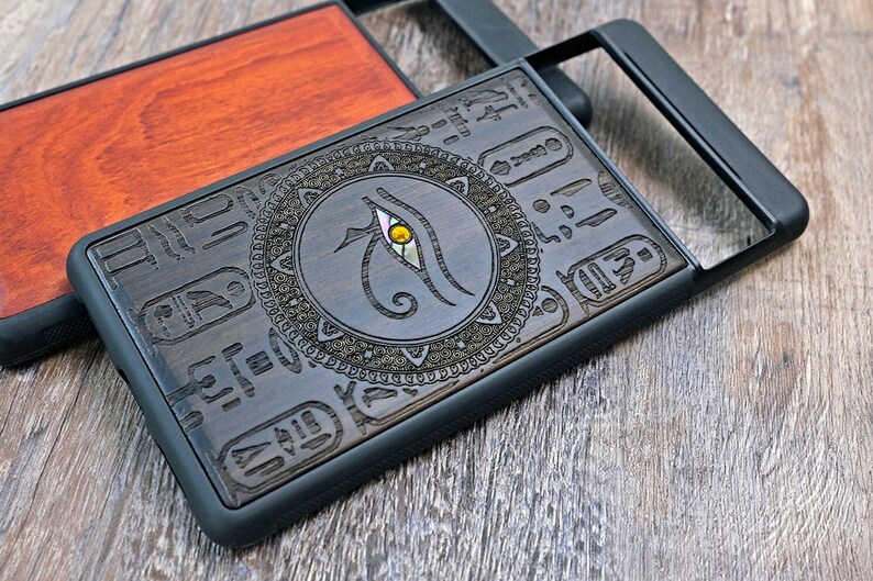 Op de afbeelding: Een zwarte telefoonhoes met een houten inlay met een Egyptisch oog van Horus-ontwerp. Het oog is omgeven door een cirkelvormig patroon en hi&euml;rogliefen.