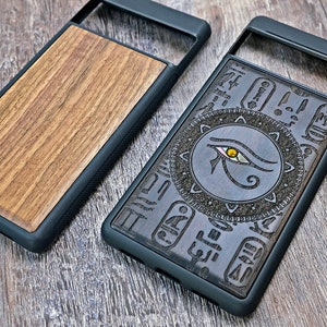Op de afbeelding: Twee telefoonhoesjes van hout met een zwarte rubberen rand. Een hoesje heeft een houtnerfpatroon en de andere heeft een ingewikkeld ontwerp met het oog van Horus en hi&euml;rogliefen.