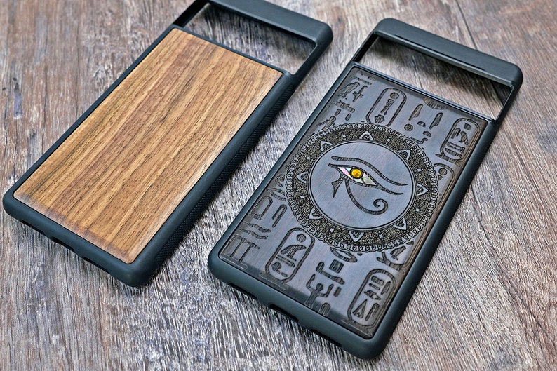 Op de afbeelding: Twee telefoonhoesjes van hout met een zwarte rubberen rand. Een hoesje heeft een houtnerfpatroon en de andere heeft een ingewikkeld ontwerp met het oog van Horus en hi&euml;rogliefen.