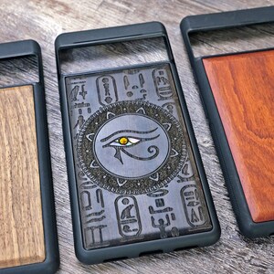 Op de afbeelding: Drie telefoonhoesjes met verschillende houtontwerpen. De eerste hoes heeft een donker houtontwerp met een Egyptisch oogsymbool. De tweede hoes heeft een licht houtontwerp met een eenvoudig nerfpatroon. De derde hoes heeft een roodbruin houtontwerp.
