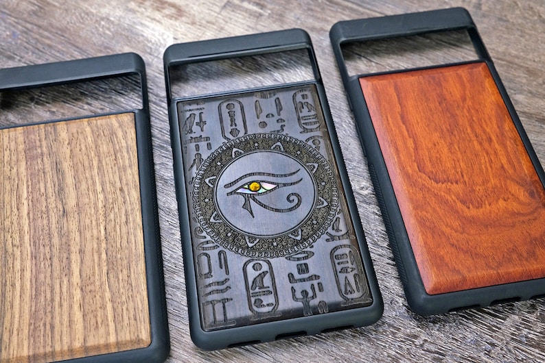 Op de afbeelding: Drie telefoonhoesjes met verschillende houtontwerpen. De eerste hoes heeft een donker houtontwerp met een Egyptisch oogsymbool. De tweede hoes heeft een licht houtontwerp met een eenvoudig nerfpatroon. De derde hoes heeft een roodbruin houtontwerp.