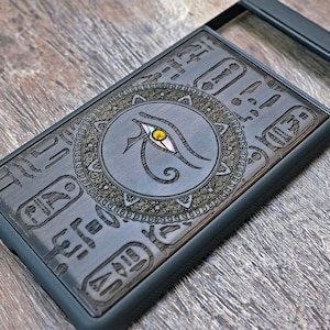 Op de afbeelding: Een zwarte telefoonhoes met een houten inlay met een Egyptisch oog van Horus-ontwerp. Het ontwerp is omgeven door een decoratieve rand en bevat hi&euml;rogliefen.