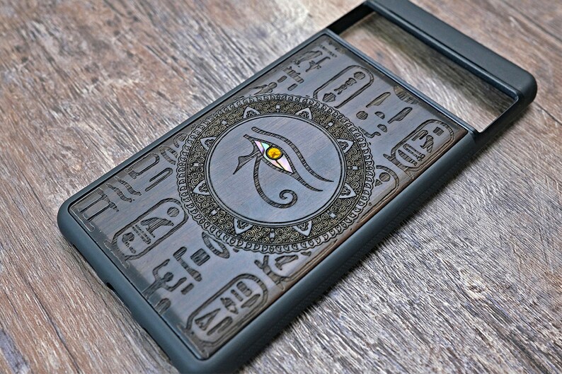 Op de afbeelding: Een zwarte telefoonhoes met een houten inlay met een Egyptisch oog van Horus-ontwerp. Het ontwerp is omgeven door een decoratieve rand en bevat hi&euml;rogliefen.