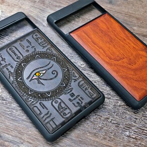 Op de afbeelding: Twee telefoonhoesjes met een zwarte rubberen rand. Een hoesje heeft een houten paneel met een gegraveerd oog van Horus-ontwerp. Het andere hoesje heeft een effen houten paneel.