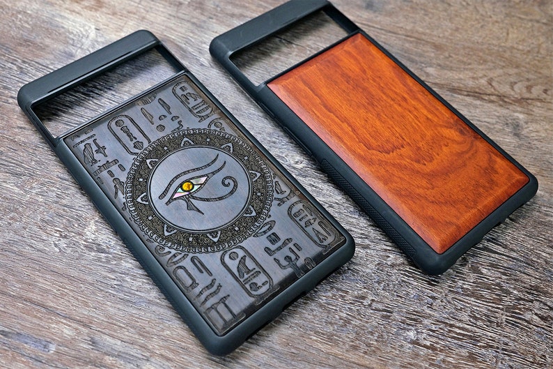 Op de afbeelding: Twee telefoonhoesjes met een zwarte rubberen rand. Een hoesje heeft een houten paneel met een gegraveerd oog van Horus-ontwerp. Het andere hoesje heeft een effen houten paneel.