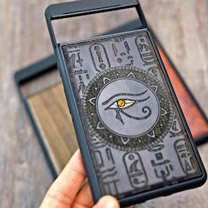 Op de afbeelding: Zwarte telefoonhoes met een transparant achterpaneel met een gegraveerd ontwerp van het oog van Horus omringd door hi&euml;rogliefen.