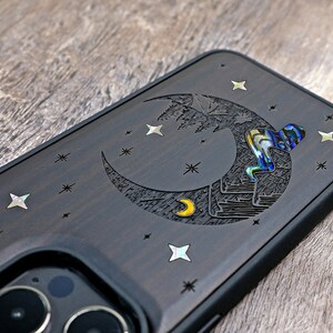 Puede incluir: Una funda de tel&eacute;fono marr&oacute;n oscuro con un dise&ntilde;o de luna creciente y estrellas. La luna est&aacute; tallada con una luna creciente m&aacute;s peque&ntilde;a y una figura que escala la luna. Las estrellas est&aacute;n hechas de material iridiscente.