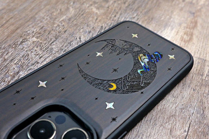 Puede incluir: Una funda de tel&eacute;fono marr&oacute;n oscuro con un dise&ntilde;o de luna creciente y estrellas. La luna est&aacute; tallada con una luna creciente m&aacute;s peque&ntilde;a y una figura que escala la luna. Las estrellas est&aacute;n hechas de material iridiscente.