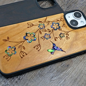 May include: A wooden phone case with a black border, featuring a hummingbird and floral design with iridescent accents. The case is made of natural wood and has a unique, handcrafted look.