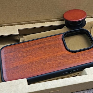 Puede incluir: Una funda de tel&eacute;fono de madera con un borde negro y un recorte para el objetivo de la c&aacute;mara. La funda est&aacute; en una caja de cart&oacute;n con un forro de fieltro marr&oacute;n.
