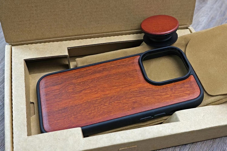 Puede incluir: Una funda de tel&eacute;fono de madera con un borde negro y un recorte para el objetivo de la c&aacute;mara. La funda est&aacute; en una caja de cart&oacute;n con un forro de fieltro marr&oacute;n.