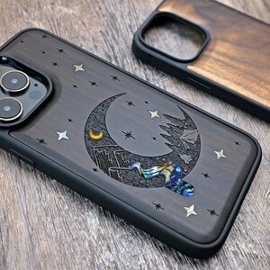 Puede incluir: Una funda de tel&eacute;fono de madera negra con un dise&ntilde;o de luna creciente, estrellas y una peque&ntilde;a pieza de n&aacute;car iridiscente. La funda est&aacute; dise&ntilde;ada para un iPhone.