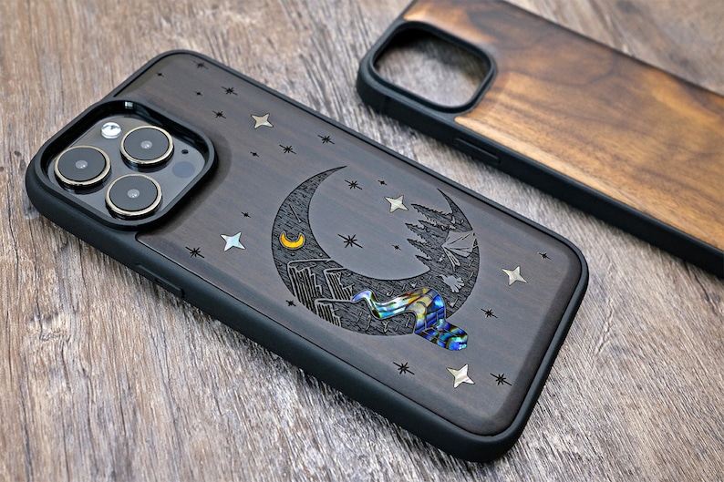 Puede incluir: Una funda de tel&eacute;fono de madera negra con un dise&ntilde;o de luna creciente, estrellas y una peque&ntilde;a pieza de n&aacute;car iridiscente. La funda est&aacute; dise&ntilde;ada para un iPhone.