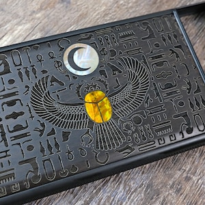 Puede incluir: Una funda rectangular negra con un diseño egipcio grabado. La funda presenta un escarabajo con un centro amarillo y una luna creciente. El diseño se encuentra sobre un fondo oscuro con jeroglíficos.