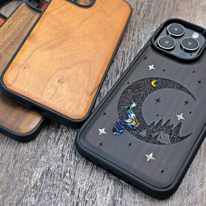 Puede incluir: Una funda de tel&eacute;fono de madera negra con un dise&ntilde;o tallado de una luna creciente, estrellas y una escena de monta&ntilde;a. La funda tiene un parachoques de goma negro y una cubierta de lente de c&aacute;mara transparente.
