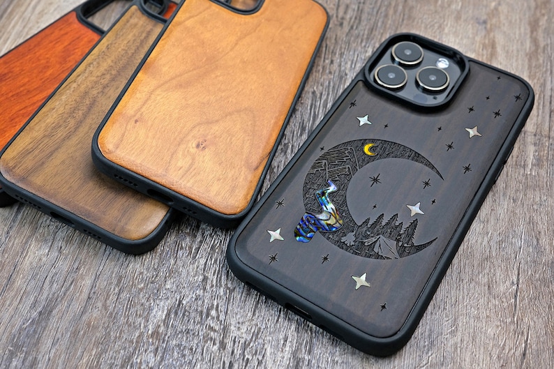 Puede incluir: Una funda de tel&eacute;fono de madera negra con un dise&ntilde;o tallado de una luna creciente, estrellas y una escena de monta&ntilde;a. La funda tiene un parachoques de goma negro y una cubierta de lente de c&aacute;mara transparente.