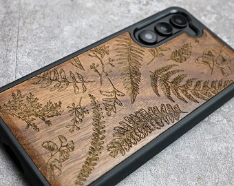 May include: A brown wooden phone case with a black rubber bumper. The case has a laser-etched design of ferns and leaves.