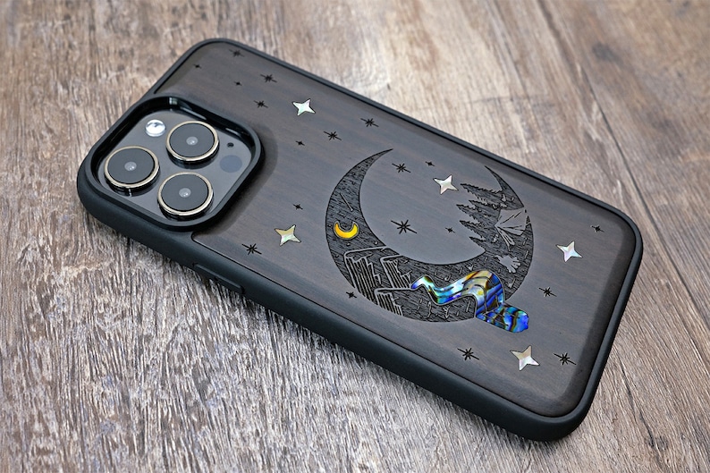 Puede incluir: Una funda para tel&eacute;fono de madera negra con un dise&ntilde;o de luna creciente y estrellas. La luna est&aacute; hecha de n&aacute;car y tiene una peque&ntilde;a luna creciente en su interior. La funda tiene un parachoques de goma negro.