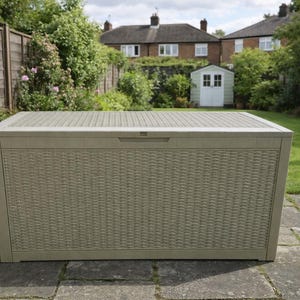 May include: A beige outdoor storage chest with a woven pattern on the front and sides. The lid has a ridged design. The chest is set on a stone patio in a garden, with a house and trees in the background.