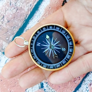 May include: A gold compass with a black face and a silver ring. The compass is marked with numbers from 0 to 360 and has a north, south, east, and west arrow.