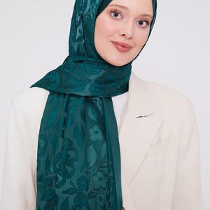 Puede incluir: Un hiyab verde azulado con un estampado floral. El velo está drapeado sobre la cabeza y los hombros. La persona lleva una blazer color crema.