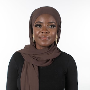 May include: A woman wearing a brown hijab and a black long-sleeve shirt. The hijab is draped around the head and shoulders. The woman is wearing gold earrings. The background is plain white.